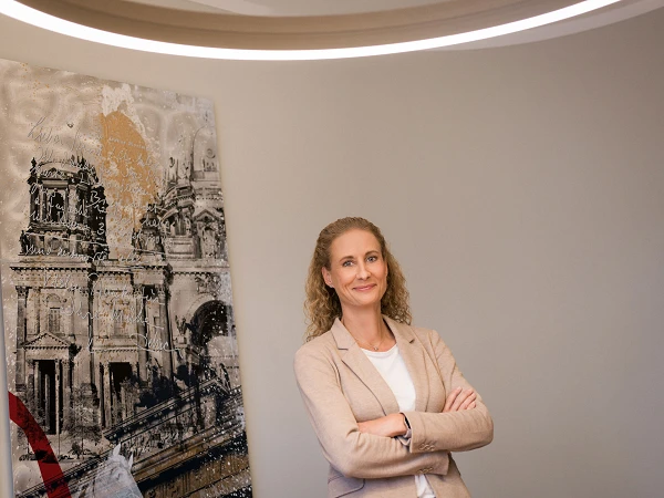 Eine Frau mit lockigem, blondem Haar steht lächelnd mit verschränkten Armen vor einer modernen Wandgestaltung, die ein künstlerisches Motiv zeigt. Sie trägt einen hellbeigen Blazer und einen weißen Oberteil. Im Hintergrund ist eine stilisierte Darstellung von Architektur zu sehen.