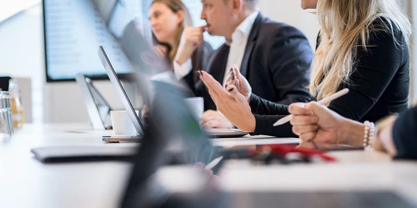 In einem modernen Besprechungsraum diskutieren mehrere Personen aktiv, während sie Laptops und Tablets nutzen. Im Vordergrund sind Hände mit einem Stift und Notizen zu sehen, während im Hintergrund eine Person am Tisch spricht. An der Wand ist ein Bildschirm mit Daten oder Diagrammen sichtbar.