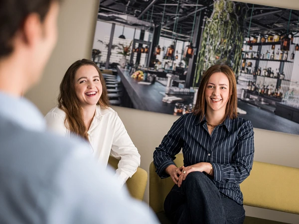 Zwei Frauen sitzen auf einem Sofa und lächeln, während sie mit einer anderen Person sprechen. Im Hintergrund ist ein großes Bild einer modernen Bar oder Lounge zu sehen.