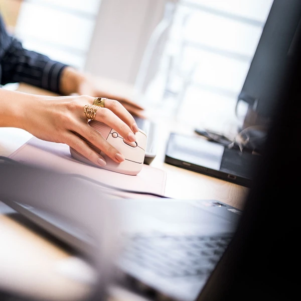 Eine Hand mit einem Ring bewegt eine Computermaus über ein Mauspad, während auf einem Schreibtisch ein Laptop und andere Bürogegenstände zu sehen sind.