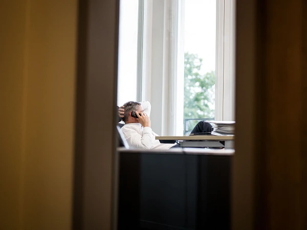 Ein Mann sitzt entspannt auf einem Bürostuhl und telefoniert, während er sich zurücklehnt und durch ein Fenster auf den Außenbereich schaut.
