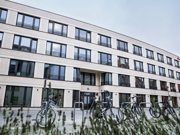 Ein modernes Gebäude mit einer abgerundeten Fassade, großen Fenstern und einer minimalistischen Außenarchitektur, davor stehen mehrere Fahrräder auf einem Stellplatz. Im Vordergrund sind Pflanzen zu sehen.
