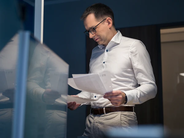Ein Mann in einem weißen Hemd steht in einem modernen Büro und betrachtet nachdenklich Dokumente in seinen Händen.