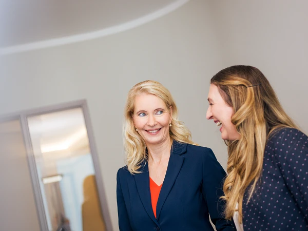 Zwei Frauen stehen in einem Büro und lächeln sich an, während sie miteinander sprechen. Die Frau links trägt einen blauen Blazer und hat blonde Haare, die Frau rechts hat lange, lockige Haare und trägt eine gepunktete Jacke.