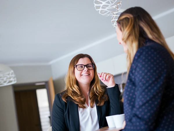 Zwei Frauen unterhalten sich an einem Tisch in einem modernen Raum, während eine von ihnen eine Tasse in der Hand hält und lächelt.