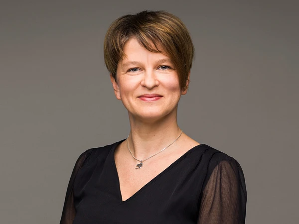 Eine lächelnde Frau mit kurzen, braunen Haaren trägt ein schwarzes Kleid mit langen, transparenten Ärmeln und steht vor einem neutralen Hintergrund.