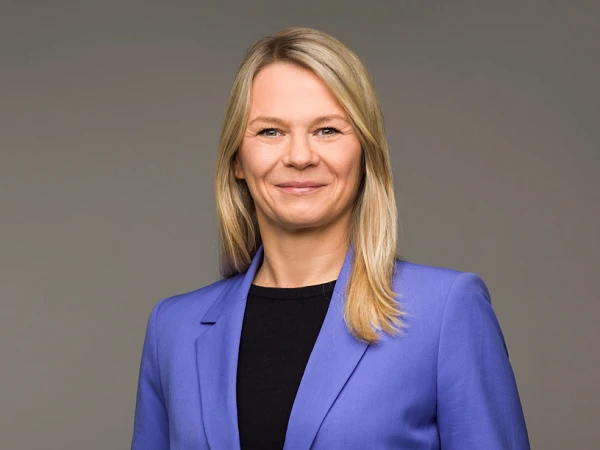 Eine lächelnde Frau mit langen, blonden Haaren trägt einen blauen Blazer und steht vor einem grauen Hintergrund.