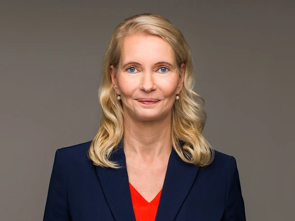 Eine lächelnde Frau mit langen, blonden Haaren trägt einen dunkelblauen Blazer über einem roten Oberteil. Sie steht vor einem grauen Hintergrund und blickt direkt in die Kamera.