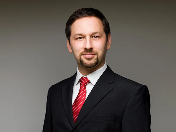 Ein Mann in einem schwarzen Anzug mit einem weißen Hemd und einer roten Krawatte steht vor einem neutralen Hintergrund und schaut direkt in die Kamera.