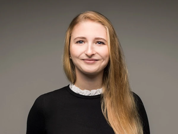 Eine Person mit langen, blonden Haaren lächelt freundlich in die Kamera, dressed in einem schwarzen Pullover mit einem weißen Kragen. Der Hintergrund ist einfarbig grau.