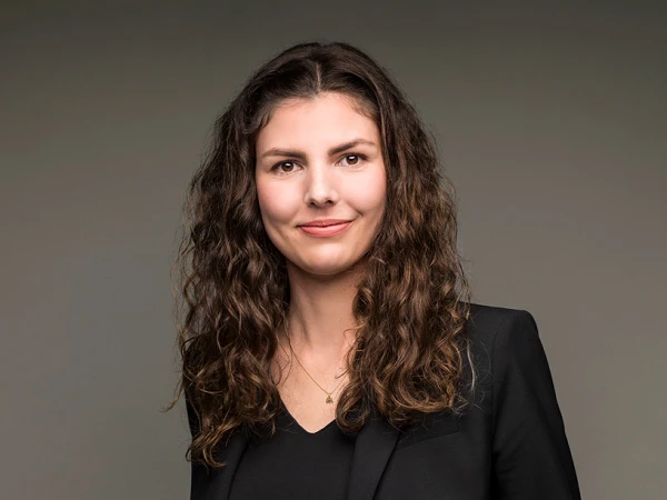Eine Frau mit lockigem, braunem Haar trägt einen schwarzen Blazer und eine schwarze Bluse sowie eine rustikale, braune Hose. Sie lächelt freundlich und steht vor einem neutralen Hintergrund.