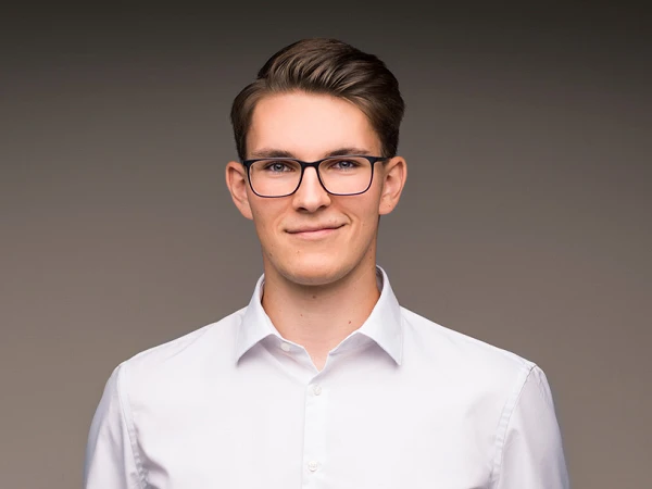 Ein junger Mann mit Brille trägt ein weißes Hemd und eine beige Hose. Er steht lächelnd vor einem neutralen, grauen Hintergrund.