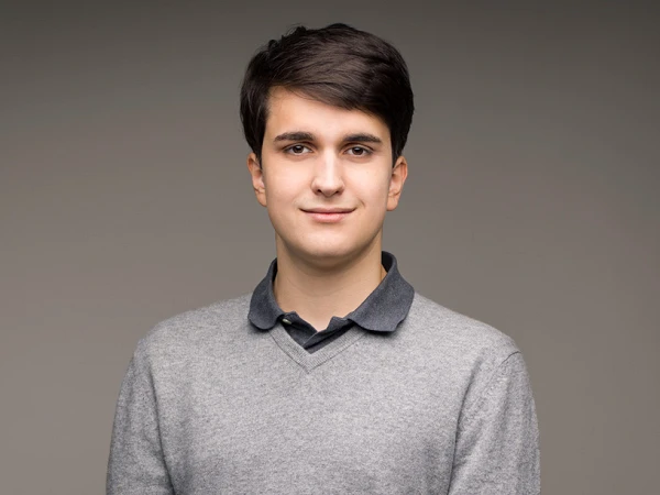 Ein junger Mann mit dunklen Haaren steht mit verschränkten Händen vor einem neutralen, grauen Hintergrund und trägt einen grauen Pullover über einem blauen Poloshirt.