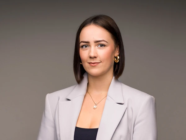Eine Person in einem grauen Blazer und schwarzem Oberteil steht mit verschränkten Armen vor einem neutralen Hintergrund. Sie hat kurzes, braunes Haar und trägt goldene Ohrringe.