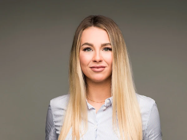Eine junge Frau mit langen, blonden Haaren trägt eine gestreifte Bluse und steht mit beiden Händen in den Taschen ihrer Hose. Sie lächelt leicht vor einem grauen Hintergrund.