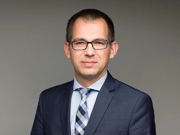 Eine geschäftlich gekleidete Person mit Brille steht aufrecht und hält die Hände vor sich, während sie in die Kamera schaut. Der Hintergrund ist neutral und glatt. Die Person trägt einen dunkelblauen Anzug und ein kariertes Hemd.