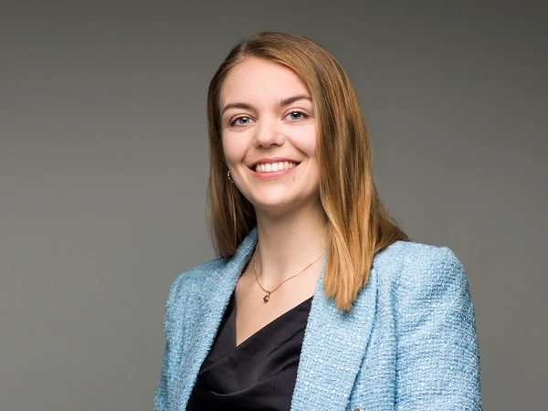 Eine smilende Frau mit langen, blonden Haaren trägt eine hellblaue Jacke über einem schwarzen Kleid und steht vor einem neutralen Hintergrund.