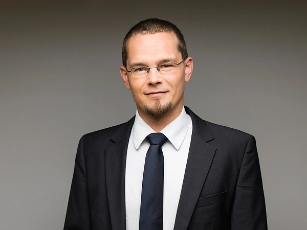 Ein Mann in einem Anzug mit einem weißen Hemd und einer dunklen Krawatte steht lässig mit einer Hand in der Hosentasche vor einem neutralen Hintergrund.