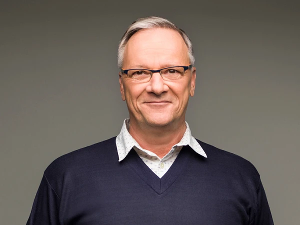 Ein älterer Mann mit Brille trägt einen dunkelblauen Pullover über einem hellen Hemd und steht mit verschränkten Händen vor einem einfarbigen Hintergrund.
