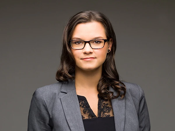 Eine selbstbewusste Frau mit schulterlangen, braunen Haaren und Brille trägt ein graues Blazer-Jackett über einem schwarzen Oberteil. Sie steht vor einem neutralen Hintergrund und schaut direkt in die Kamera.