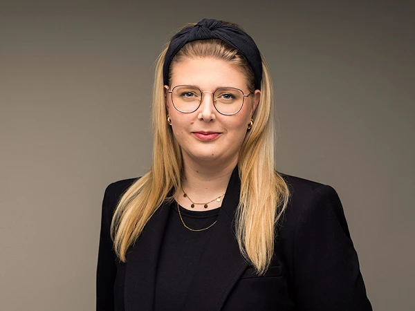 Eine Frau mit langen blonden Haaren und einer schwarzen Bluse trägt einen eleganten schwarzen Blazer. Sie hat eine modische Brille auf und steht mit verschränkten Händen vor einem neutralen Hintergrund.