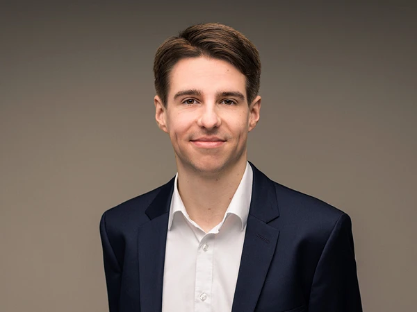 Ein junger Mann steht in einem dunklen Anzug mit einem weißen Hemd, mit verschränkten Armen und einem neutralen Lächeln vor einem sanft gefärbten Hintergrund.