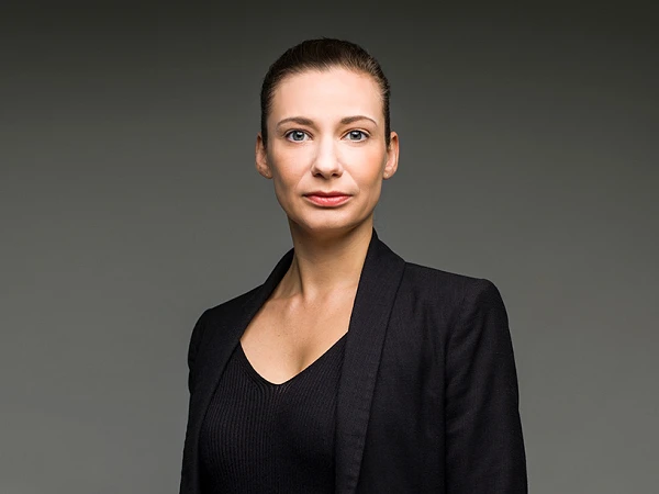 Eine Frau mit langen, glatten Haaren steht vor einem grauen Hintergrund und trägt einen schwarzen Blazer über einem schwarzen Oberteil sowie braunen Hosen. Sie schaut direkt in die Kamera und hat einen neutralen Gesichtsausdruck.