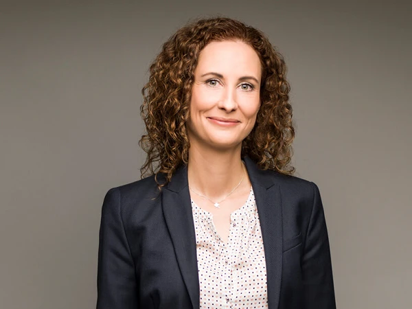 Eine Frau mit lockigem, braunem Haar trägt einen dunkelblauen Blazer über einer blassbeigen Bluse mit kleinen Punkten und lächelt in die Kamera. Der Hintergrund ist neutral und hell.