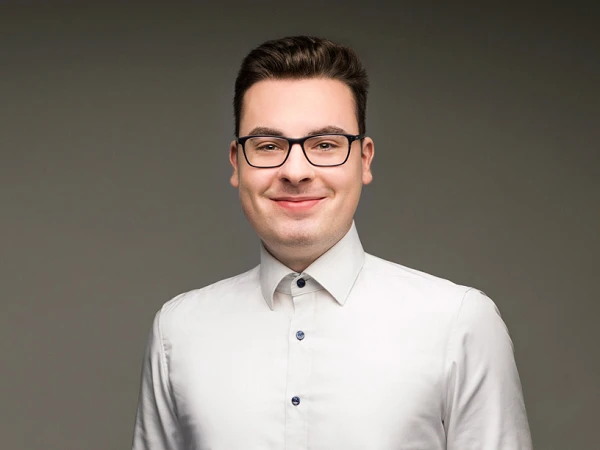 Ein junger Mann mit Brille trägt ein weißes Hemd und dunkle Hose, steht mit verschränkten Armen vor einem grauen Hintergrund und lächelt freundlich.