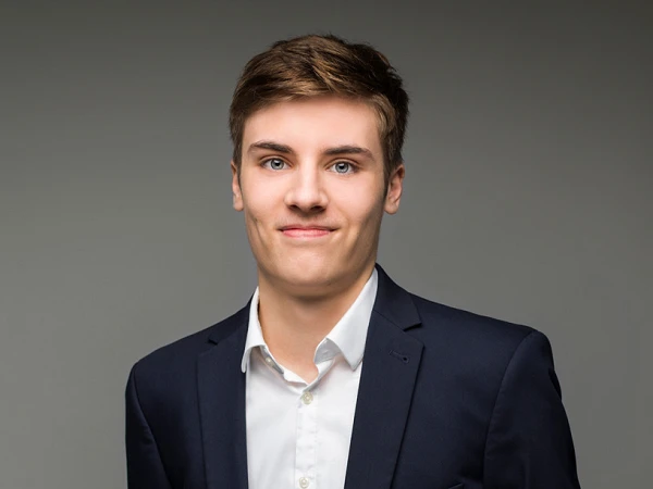 Junger Mann mit kurzer, blonder Haarfrisur trägt einen dunkelblauen Anzug und ein weißes Hemd, während er vor einem grauen Hintergrund steht.