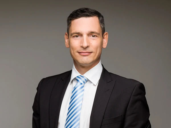 Ein Mann in einem schwarzen Anzug mit einem weißen Hemd und einer gestreiften Krawatte steht mit verschränkten Händen vor einem neutralen Hintergrund und blickt direkt in die Kamera.