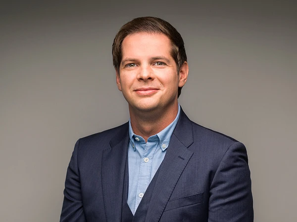Ein Mann in einem dunklen Anzug und einer hellen Hemdbluse steht mit den Händen vor sich verschränkt vor einem neutralen Hintergrund.