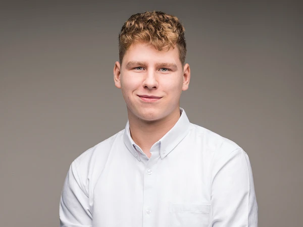 Ein junger Mann mit kurzen, lockigen Haaren trägt ein weißes Hemd und schaut freundlich in die Kamera, während er seine Hände vor dem Bauch faltet. Der Hintergrund ist einheitlich grau.