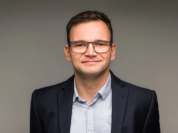Ein freundlich lächelnder Mann trägt ein dunkelblaues Sakko über einem hellblauen Hemd und steht mit den Händen hinter dem Rücken vor einem neutralen Hintergrund.