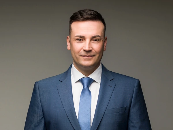 Ein Mann in einem eleganten blauen Anzug mit einem hellblauen Krawatte steht vor einem neutralen Hintergrund und schaut direkt in die Kamera.