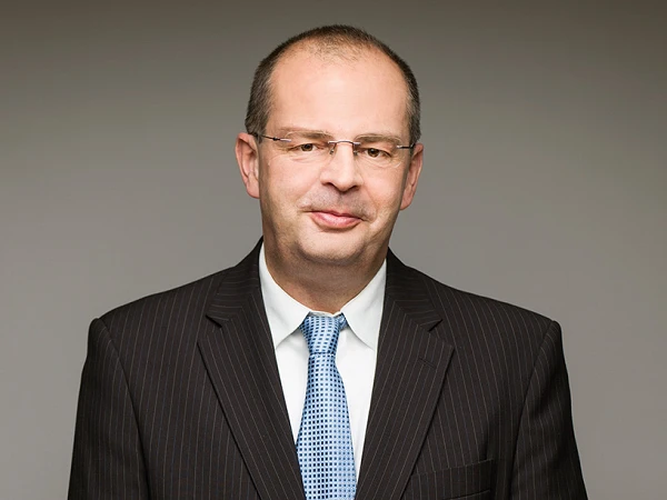 Ein Mann steht in einem formellen, gestreiften Anzug mit Krawatte und lächelt leicht in die Kamera. Der Hintergrund ist neutral und einfarbig.