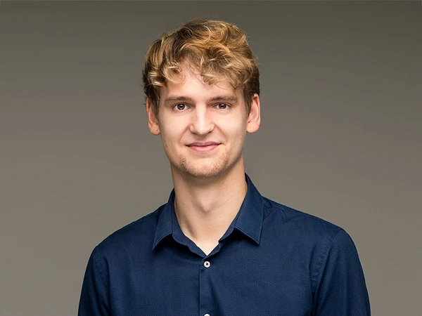 Ein junger Mann mit blonden lockigen Haaren trägt ein dunkelblaues Hemd und steht lächelnd vor einem grauen Hintergrund.