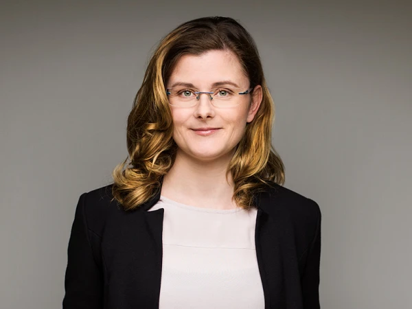 Eine selbstbewusste Frau mit langen, welligen Haaren trägt eine schwarze Jacke über einem hellen Oberteil und blickt direkt in die Kamera. Der Hintergrund ist neutral und hebt ihre Präsenz hervor.