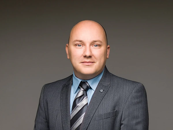 Ein Mann in einem grauen Anzug mit blauer Hemd und gestreifter Krawatte steht mit verschränkten Händen vor einem neutralen Hintergrund.