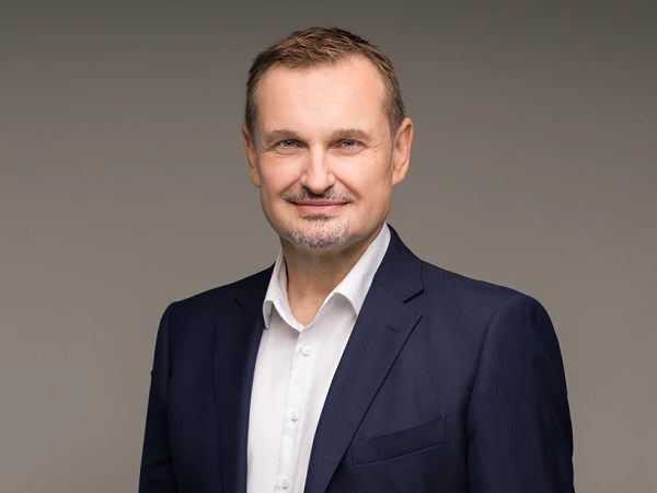 Ein mittelalterlicher Mann in einem dunklen Anzug und einem weißen Hemd steht aufrecht vor einem neutralen Hintergrund, lächelt freundlich und hat eine gepflegte Erscheinung.