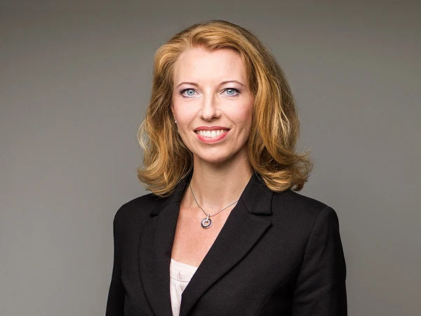 Eine Frau mit langen, blonden Haaren trägt einen schwarzen Blazer und steht in einer neutralen Pose vor einem einfarbigen Hintergrund. Sie lächelt freundlich in die Kamera.