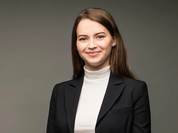Eine junge Frau in einem schwarzen Blazer und weißem Rollkragenpullover steht lächelnd mit verschränkten Händen vor einem einfarbigen Hintergrund.