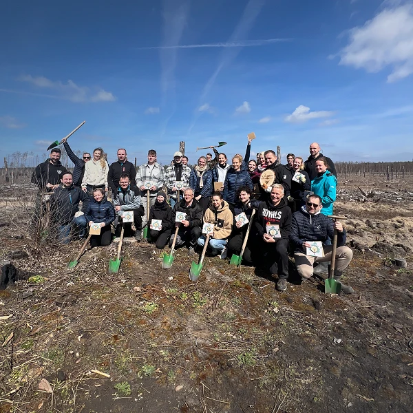 Gruppenshot von 24 Personen in einem kahlen Waldgebiet, alle halten Schaufeln und stehen in einer Pose des Feierns. Im Hintergrund sind teilweise abgeholzte Bäume und ein blauer Himmel mit wolkigen Abschnitten zu sehen.
