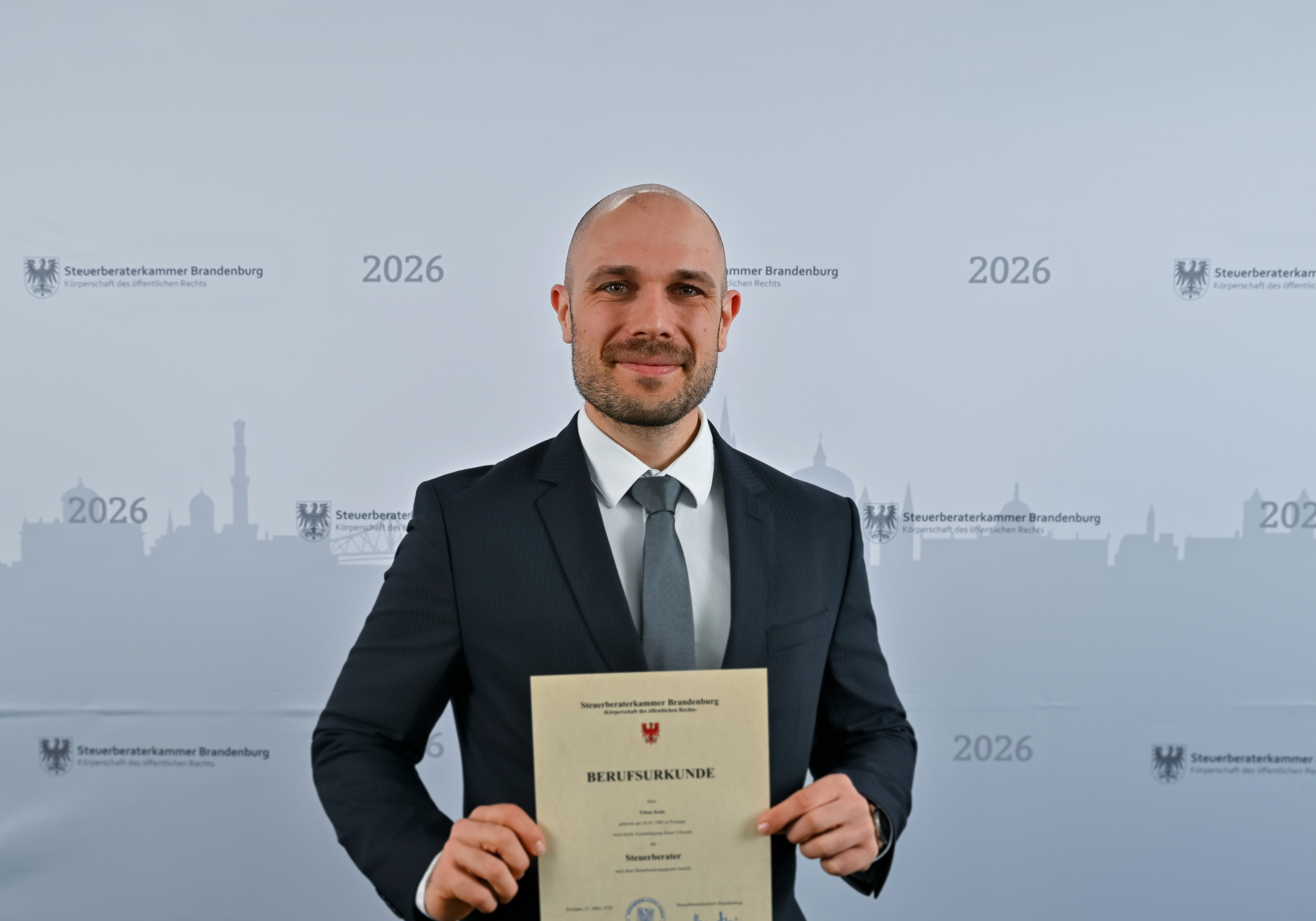 Ein Mann in einem dunklen Anzug hält stolz ein Dokument mit der Beschriftung „BERUFSBILDERUNGE“ und posiert vor einer grafischen Hintergrunddarstellung einer Stadtlandschaft mit dem Jahr 2026.