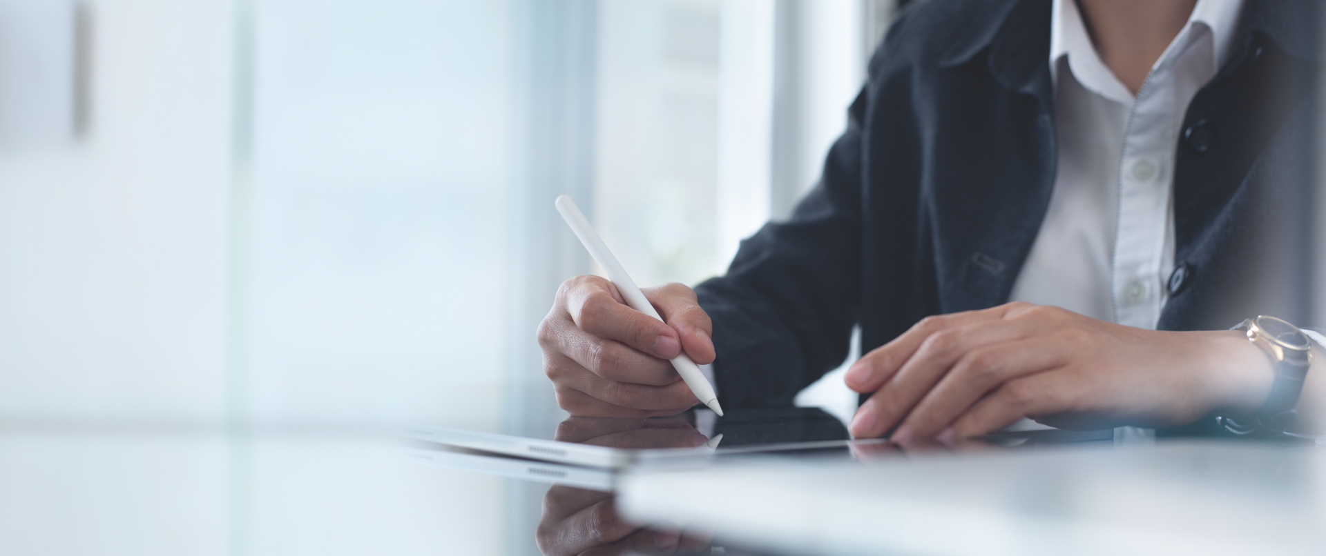 Eine Person sitzt an einem Tisch und schreibt mit einem Stylus auf einem Tablet, während sie eine elegante, schwarze Jacke und ein weißes Hemd trägt.