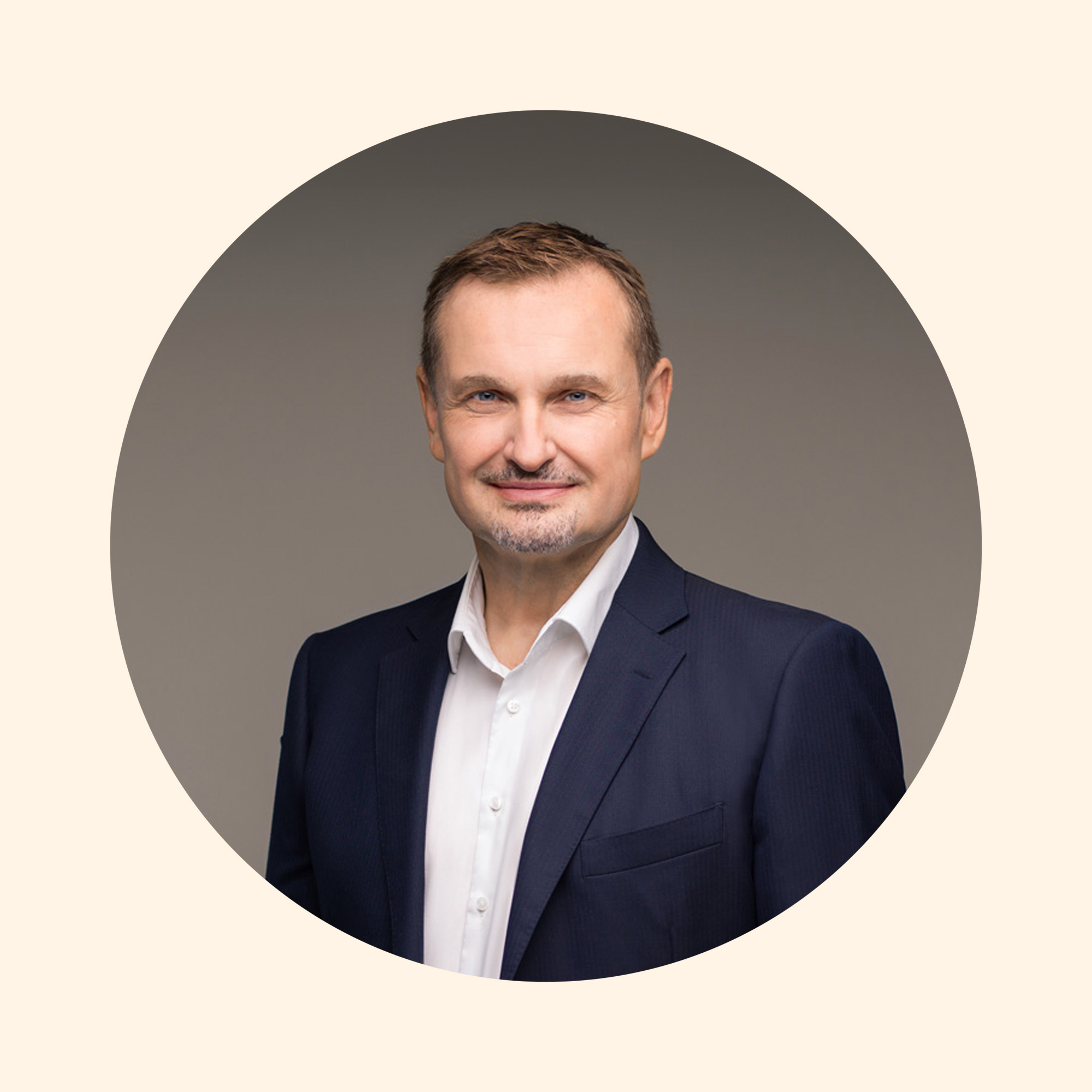 Ein Mann mit kurzen, dunklen Haaren und einem gepflegten Bart trägt einen dunklen Anzug und ein helles Hemd. Er steht vor einem neutralen Hintergrund und lächelt leicht.