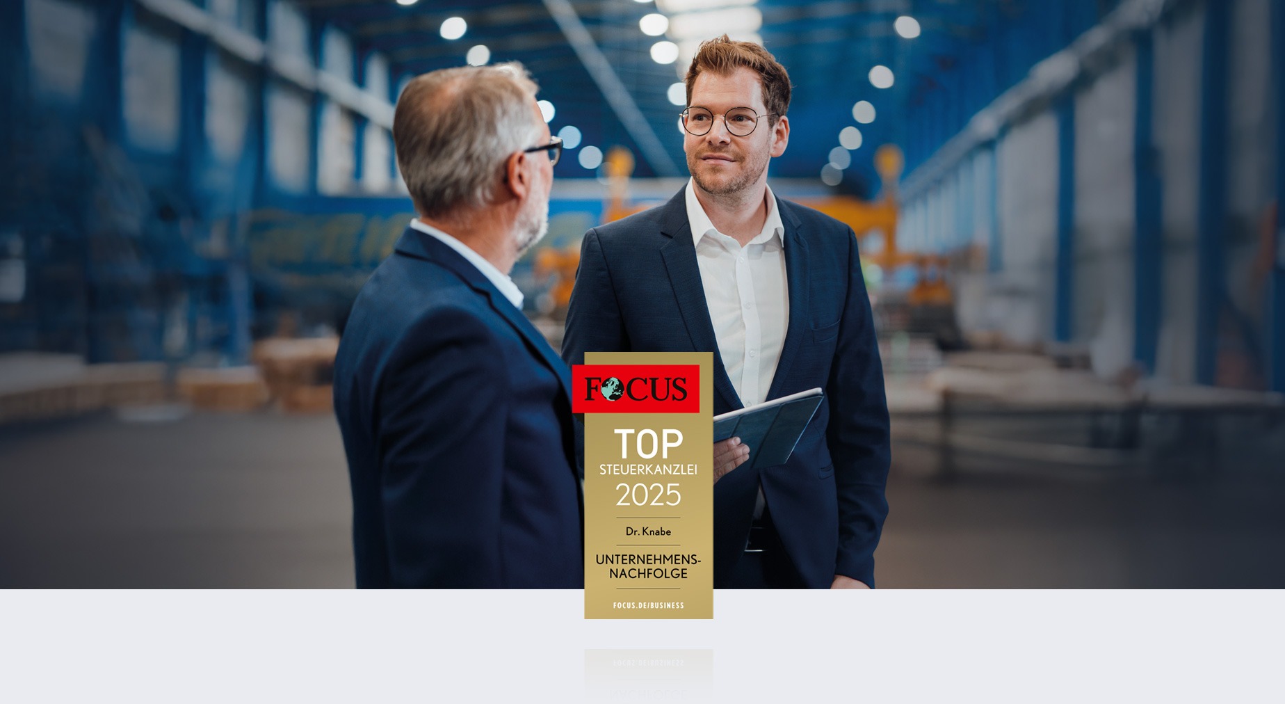 Zwei Männer stehen in einer Lagerhalle und sprechen miteinander, während einer von ihnen ein Tablet in der Hand hält. Im Vordergrund ist ein Zertifikat mit der Aufschrift „FOCUS TOP STEUERKANDEL 2025“ abgebildet.