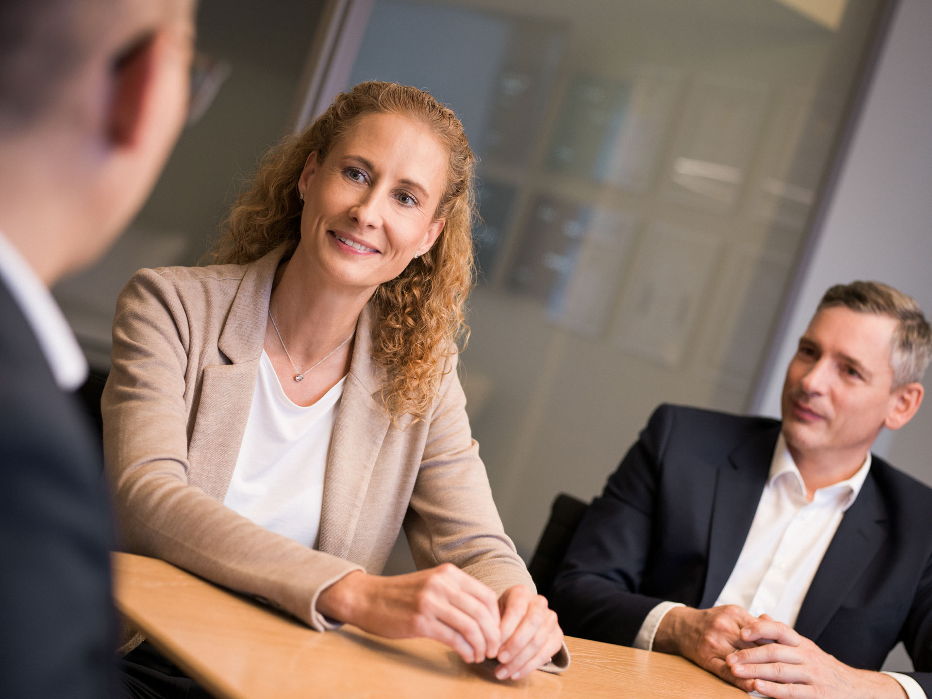 Eine Frau mit lockigem, blonde Haar sitzt an einem Tisch in einem Konferenzraum und lächelt, während sie mit zwei weiteren Personen spricht, darunter ein Mann im Anzug und eine weitere Person, die teilweise im Bild sichtbar ist.