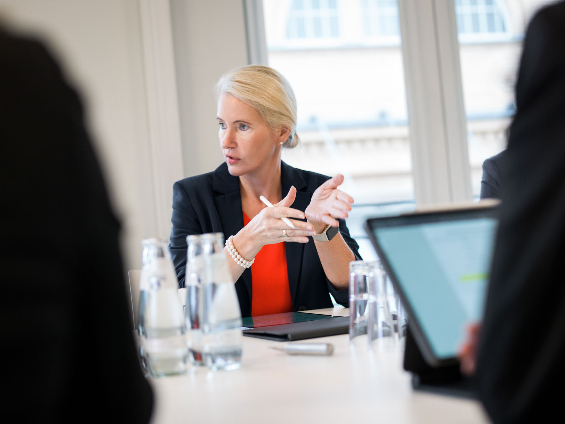 Eine Frau in einem schwarzen Blazer und rotem Oberteil spricht engagiert während eines Meetings, während mehrere Wassergläser und ein Laptop auf dem Tisch vor ihr stehen.
