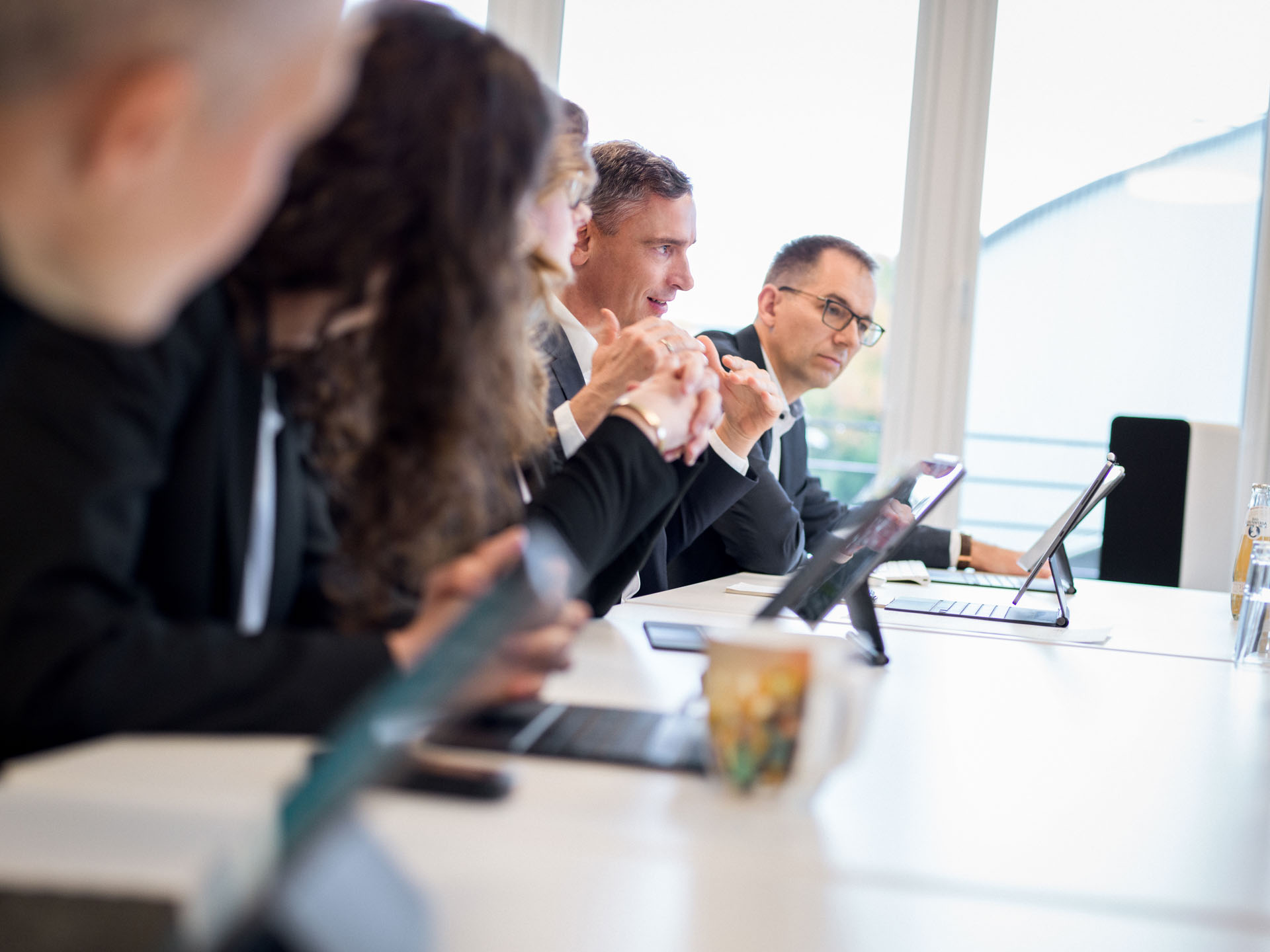 Eine Gruppe von fünf Personen in formeller Kleidung sitzt an einem Tisch und schaut auf Tablets, während sie an einer Besprechung teilnehmen. Im Hintergrund sind Fenster sichtbar, die Tageslicht hereinlassen.